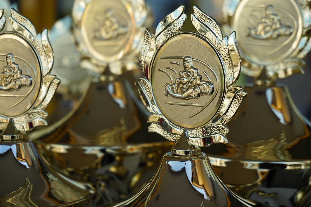 A close-up shot shows a hand holding a small, golden trophy with a star on top, against a blurred background of other similar trophies.