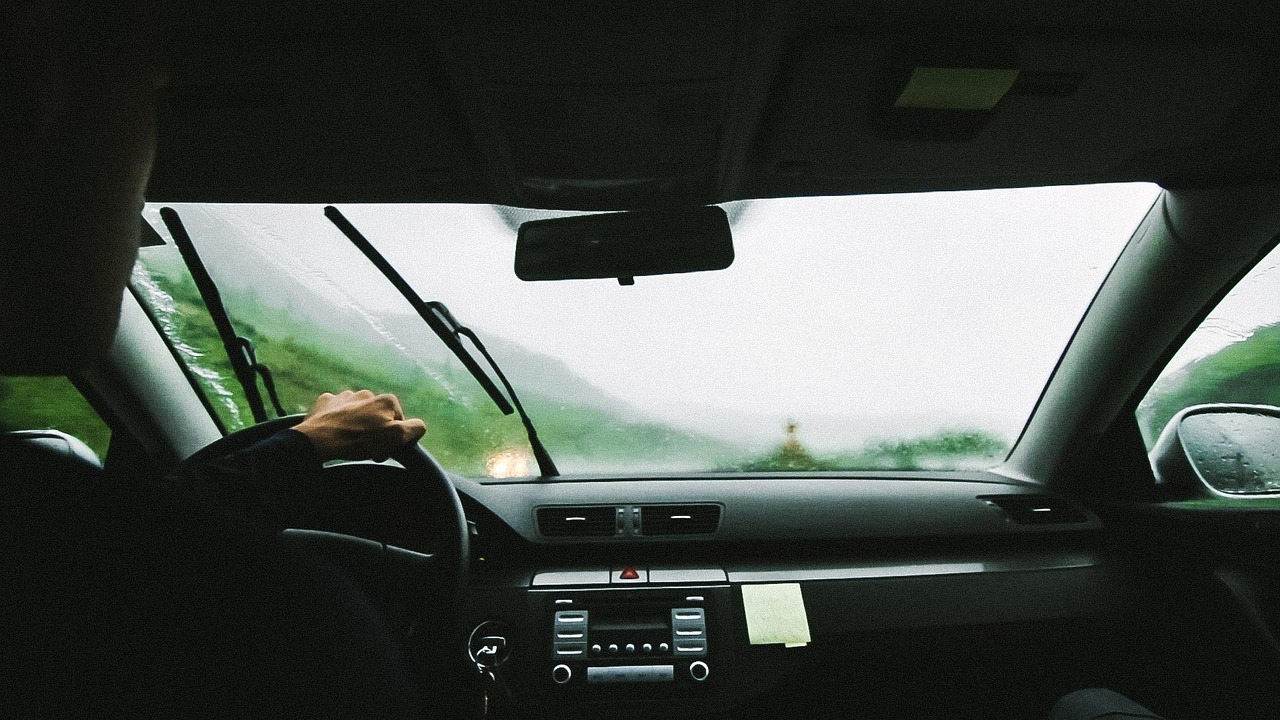 A close-up shot shows a car's windshield with visible scratches and streaks, likely caused by windshield wipers, with a blurred background.