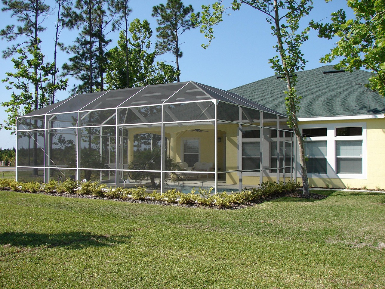 A modern glass conservatory or winter garden with a wooden frame is shown from the outside, featuring a glass roof and large windows, surrounded by green trees and a blue sky.